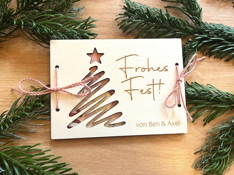 May include: A wooden Christmas card with a cut-out Christmas tree design and a star at the top. The card is tied with pink twine and has the words "Frohes Fest!" and "von Ben & Axel" engraved on it. Green pine branches surround the card.