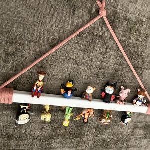Op de afbeelding: Een wit houten plank met een roze touwhouder bevat een verzameling kleine beeldjes. De beeldjes omvatten een cowboy, een vogel, een aap, een vampier, een varken, een kat, een ridder, een zeemeermin en een prinses.