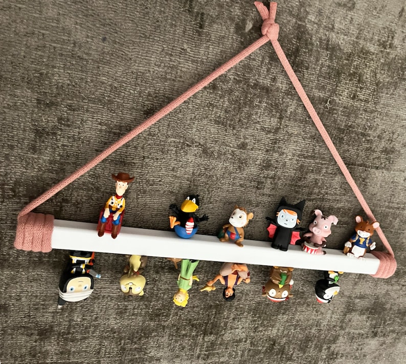 Op de afbeelding: Een wit houten plank met een roze touwhouder bevat een verzameling kleine beeldjes. De beeldjes omvatten een cowboy, een vogel, een aap, een vampier, een varken, een kat, een ridder, een zeemeermin en een prinses.