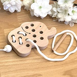 Könnte beinhalten: Holzspielzeug in Form eines Elefanten mit Löchern zum Einfädeln einer weißen Schnur. Das Spielzeug hat eine kleine Holzperle am Ende der Schnur.