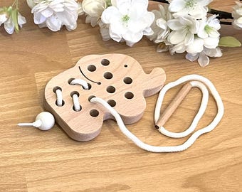 Könnte beinhalten: Holzspielzeug in Form eines Elefanten mit Löchern zum Einfädeln einer weißen Schnur. Das Spielzeug hat eine kleine Holzperle am Ende der Schnur.