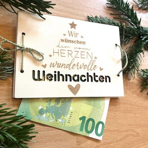May include: A wooden Christmas card with the German text "Wir w&uuml;nschen den vom Herzen wundervolle Weihnachten" is tied with twine. A 100 Euro bill is partially visible beneath the card. Green pine branches are in the background.