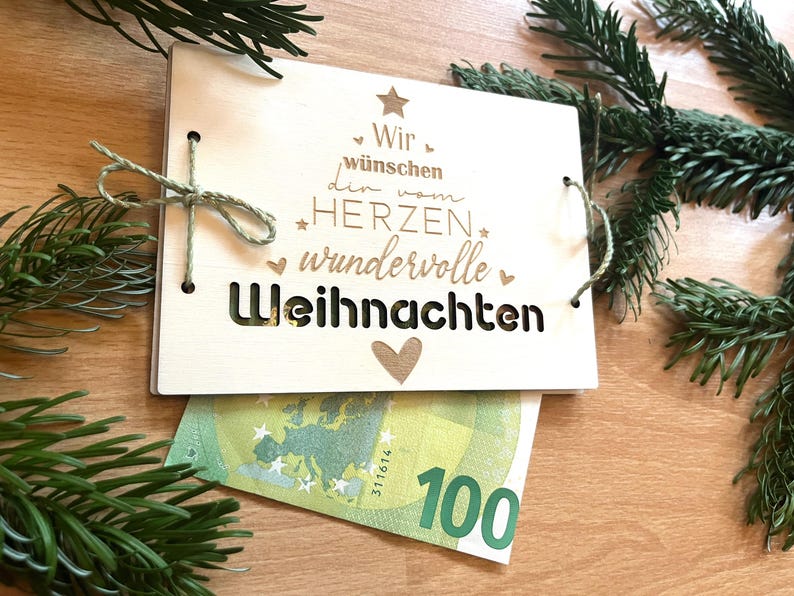 May include: A wooden Christmas card with the German text "Wir w&uuml;nschen den vom Herzen wundervolle Weihnachten" is tied with twine. A 100 Euro bill is partially visible beneath the card. Green pine branches are in the background.