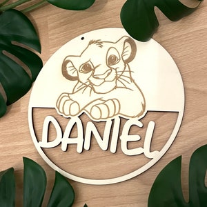Könnte beinhalten: Ein rundes Holzschild mit einem Cartoon-Bild eines Löwenjungen und dem Namen "DANIEL" in einer verspielten Schrift.