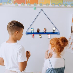 May include: Two children stand in front of a white wall with a blue wooden shelf hanging from a rope. The shelf has five small figurines on it. The figurines are of various animals and characters. The shelf is decorated with a blue and white rope.