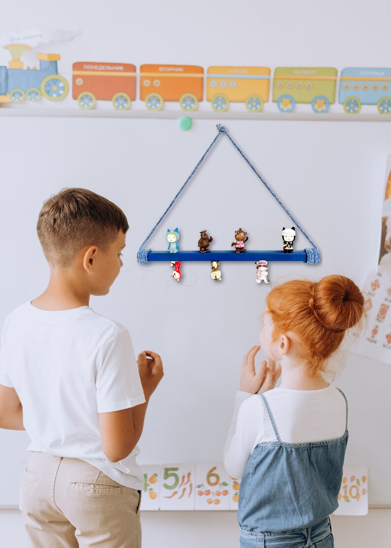 May include: Two children stand in front of a white wall with a blue wooden shelf hanging from a rope. The shelf has five small figurines on it. The figurines are of various animals and characters. The shelf is decorated with a blue and white rope.
