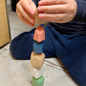 Könnte beinhalten: Ein Kind stapelt bunte geometrische Holzblöcke zu einem Turm. Die Blöcke sind in den Farben Grün, Weiß, Braun, Blau und Rosa.