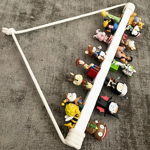 Op de afbeelding: Een wit houten plank met een touw eraan hangend. De plank is gevuld met verschillende kleine beeldjes, waaronder een bij, een varken, een cowboy, een pinguin en een vampier.