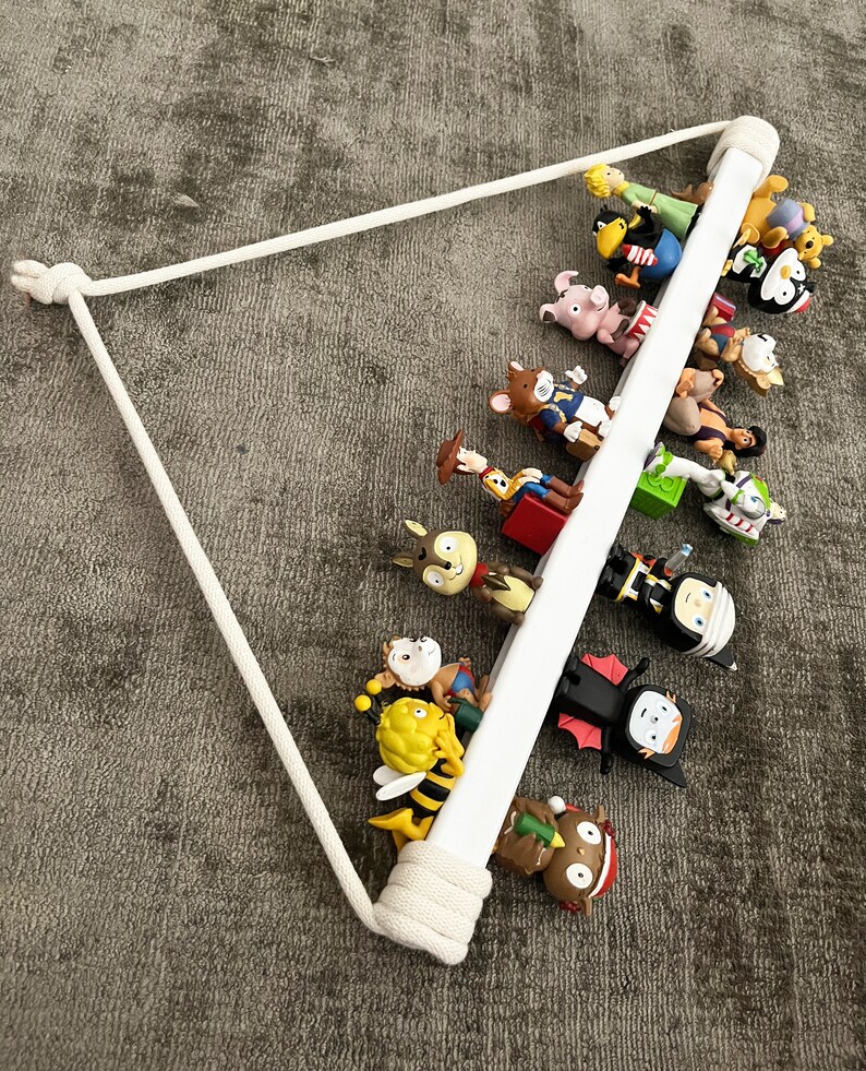 Op de afbeelding: Een wit houten plank met een touw eraan hangend. De plank is gevuld met verschillende kleine beeldjes, waaronder een bij, een varken, een cowboy, een pinguin en een vampier.