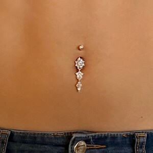 May include: A silver belly button ring with a dangling charm featuring three flower-shaped clusters of clear rhinestones.
