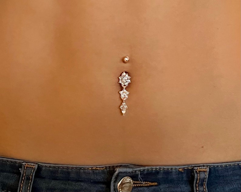 May include: A silver belly button ring with a dangling charm featuring three flower-shaped clusters of clear rhinestones.