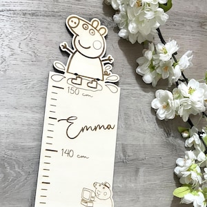 Może przedstawiać: Biała drewniana miara wzrostu z postacią kreskówkowego świniaka na górze. Wykres ma linijkę z pomiarami w centymetrach, a imię "Emma" jest napisane kursywą pod oznaczeniem 150 cm.