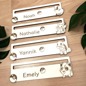 Könnte beinhalten: Vier Holz-Namensschilder mit Pokémon-Charakteren. Jedes Schild hat einen Pokéball und einen Namen: Noah, Nathalie, Yannik und Emely.