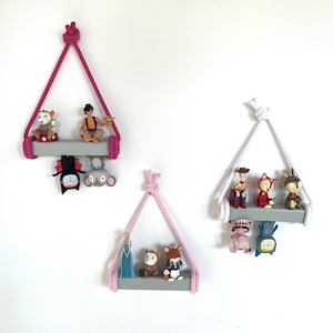 May include: Three pink and white macrame hanging shelves with grey wooden shelves. The shelves are decorated with small figurines, including a cartoon cat, a cartoon dog, a cartoon fox, a cartoon mouse, a cartoon genie, a cartoon cowboy, and a cartoon princess.