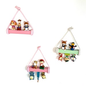 May include: Three pink and green rope shelves with wooden planks, each holding a variety of small cartoon figurines. The shelves are hung on a white wall.