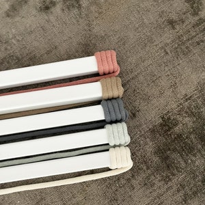 Op de afbeelding: Een set van zes koorden in verschillende kleuren, waaronder roze, beige, grijs en wit, zijn om een witte balk gewikkeld. De koorden zijn in een stapel gerangschikt, met het roze koord bovenaan en het witte koord onderaan.