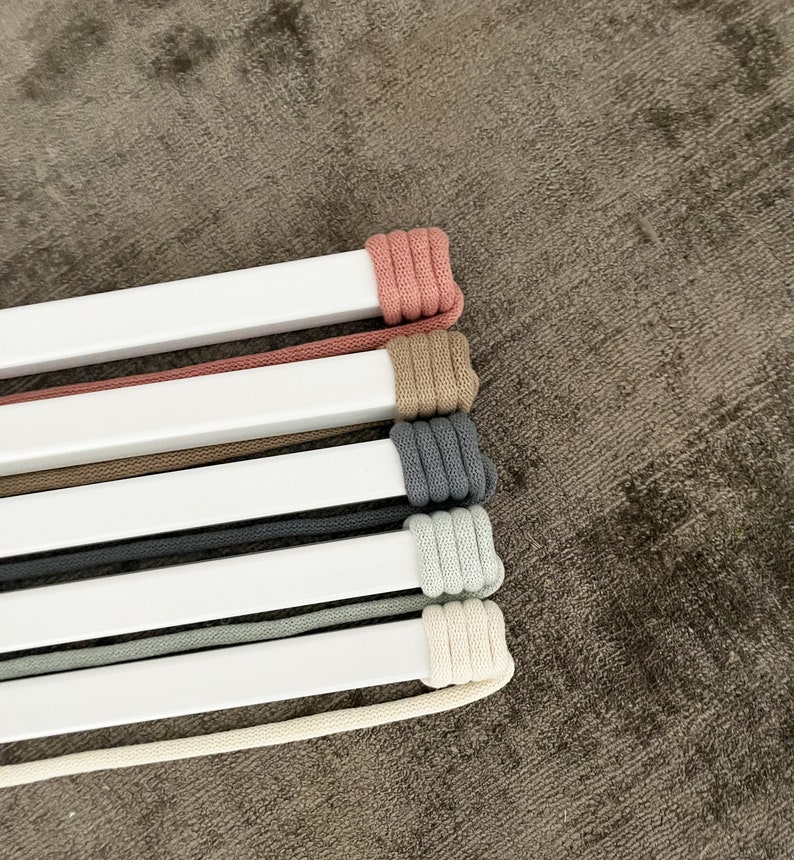 Op de afbeelding: Een set van zes koorden in verschillende kleuren, waaronder roze, beige, grijs en wit, zijn om een witte balk gewikkeld. De koorden zijn in een stapel gerangschikt, met het roze koord bovenaan en het witte koord onderaan.