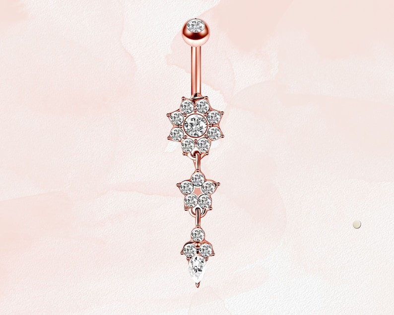 May include: A rose gold belly button ring with three dangling charms. Each charm is a flower shape with clear sparkling stones.