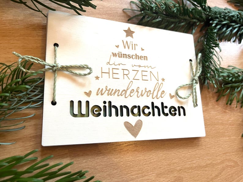 May include: A light-colored wooden Christmas card with the German text "Wir w&uuml;nschen dir vom HERZEN wundervolle Weihnachten" (We wish you a wonderful Christmas from the heart). The card is tied with green twine and decorated with a star and heart.