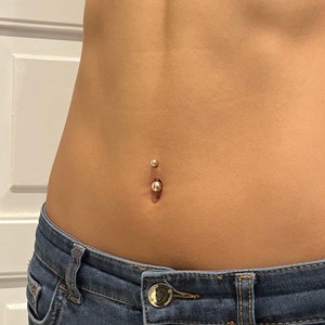Puede incluir: Primer plano de un piercing en el ombligo. El piercing presenta una pequeña bola brillante de color dorado en la parte inferior y una bola más pequeña en la parte superior. La piel es lisa y bronceada, y el piercing está centrado. Se ven unos vaqueros azules en la parte inferior.