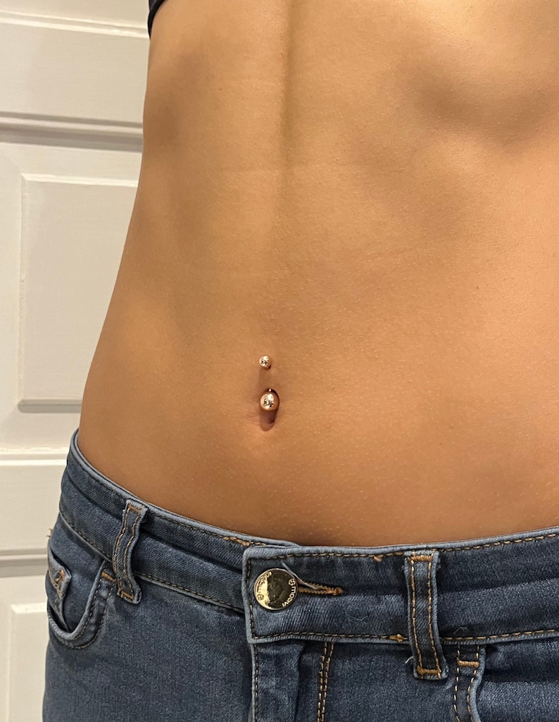 May include: A close-up of a belly button piercing. The piercing features a small, shiny, gold-colored ball on the bottom and a smaller ball on top. The skin is smooth and tan, and the piercing is centered. Blue denim jeans are visible at the bottom of the image.