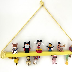 May include: A yellow wooden shelf with a rope hanger, featuring various cartoon character figurines, including Mickey Mouse, a bat, a bird, a genie, and a cowboy. The figurines are hanging from the shelf, creating a whimsical display.
