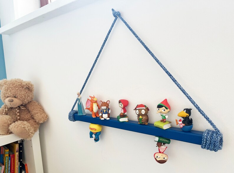 May include: A blue wooden shelf with rope hangers is mounted on a white wall. The shelf is decorated with small figurines, including a dragon, a cat, a girl, a red-hooded figure, an owl, an elf, and a black bird.