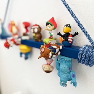 May include: A blue wooden shelf with a rope hanger, decorated with various colourful cartoon figurines. The figurines include a red-hatted gnome, a brown mouse, a blue monster, and a black bird.