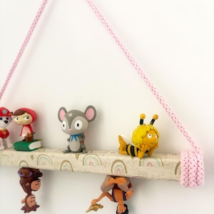 May include: A hanging shelf with a pink rope, displaying various figurines. The shelf is decorated with rainbow patterns. Figurines include a mouse, a bee, a monkey, and a character from a cartoon. The shelf is suspended against a white wall.
