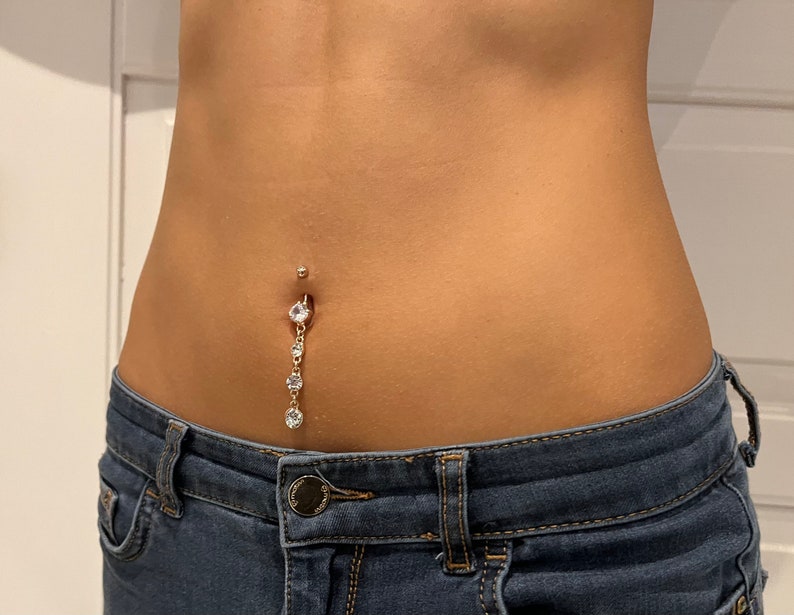 K&ouml;nnte beinhalten: Nahaufnahme des Bauches einer Person mit einem Bauchnabelpiercing, das mit einer herabh&auml;ngenden Kette aus klaren Strasssteinen verziert ist.