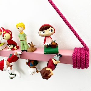 May include: A pink wooden shelf with a pink rope hanging from the ceiling. The shelf is decorated with various small figurines, including a girl in a red hood, a boy with blonde hair, a fox, and a penguin.