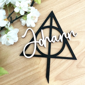 Puede incluir: Topper de pastel de madera negro en forma de triángulo con un círculo en el interior. La palabra "Johann" está escrita en cursiva blanca en el topper.