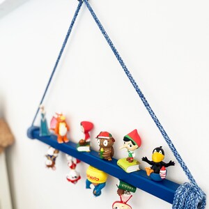 May include: A blue wooden shelf with a rope hanger, displaying a collection of small figurines. The figurines include a princess, a dragon, a girl in a red hood, an owl, a gnome, a black bird, and a few other characters. The shelf is decorated with a blue and white rope.