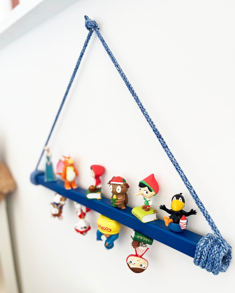 May include: A blue wooden shelf with a rope hanger, displaying a collection of small figurines. The figurines include a princess, a dragon, a girl in a red hood, an owl, a gnome, a black bird, and a few other characters. The shelf is decorated with a blue and white rope.