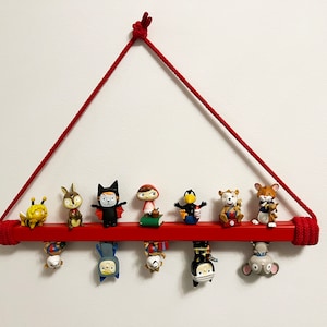 May include: A red rope wall hanging with a red wooden shelf. The shelf is decorated with small figurines of various characters, including a bee, a bat, a girl in a red hood, a bird, a tiger, and a mouse.