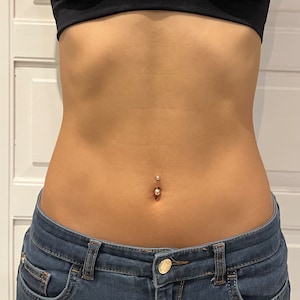 May include: Close-up of a navel piercing with a small, shiny ball on the bottom and a smaller, matching ball on top. The skin is smooth and tan, with a slight curve. The person is wearing a black top and blue jeans.