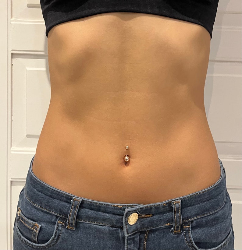 May include: Close-up of a navel piercing with a small, shiny ball on the bottom and a smaller, matching ball on top. The skin is smooth and tan, with a slight curve. The person is wearing a black top and blue jeans.