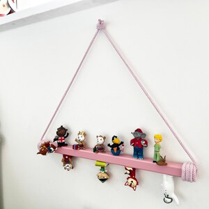 May include: A pink wooden shelf with a rope hanger, holding a collection of small figurines. The figurines include a monkey, a bear, a bird, an elephant, and a boy.