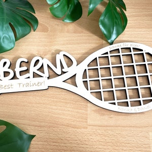 Könnte beinhalten: Ein weißes Holzschild in Form eines Tennisschlägers mit dem Namen "BERND" in großen Buchstaben. Darunter steht "Best Trainer!". Der Schläger hat ein Gittermuster und der Text "Danke für alles!" und "Norika & Daniel" sind ebenfalls sichtbar.