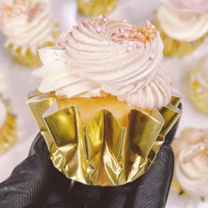 Pode incluir: Um único cupcake com cobertura de baunilha e granulado rosa e dourado, em um forro de cupcake de papel alumínio dourado. O cupcake está sendo segurado por uma mão enluvada.