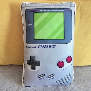 May include: A gray and white Game Boy pillow with a black and white button, a red and white button, and the text "Nintendo Game Boy" printed on the front.