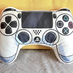 May include: A white PlayStation controller shaped pillow. The pillow has a black d-pad, buttons, and analog sticks. The PlayStation logo is in the center of the pillow.