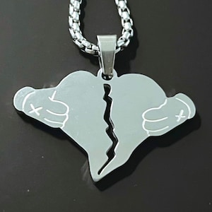 Könnte beinhalten: Eine silberne Halskette mit einem Anhänger in Form eines gebrochenen Herzens. Das Herz ist in zwei Teile geteilt, wobei jede Hälfte eine Hand mit einem 'X' auf der Rückseite zeigt.