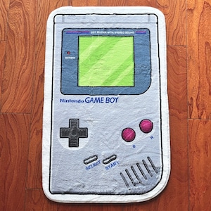 May include: A white and grey Game Boy rug with a green screen and the text "Nintendo GAME BOY" and "DOT MATRIX WITH STEREO SOUND".