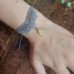 May include: A grey macrame bracelet with a decorative design and a gold-coloured charm. The bracelet is handmade with intricate knotting and features a small, leaf-shaped charm. The bracelet is shown on a person's wrist.