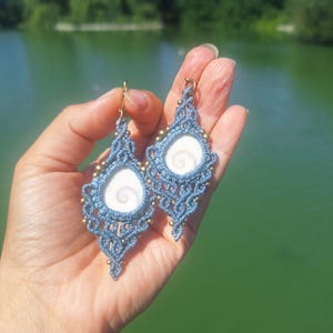 Op de afbeelding: Een paar blauwe macramé oorbellen met een centrale witte shiva-schelp met een spiraalpatroon. Het ingewikkelde ontwerp van geknoopt koord is geaccentueerd met kleine metalen kralen en afgewerkt met goudkleurige metalen haken.