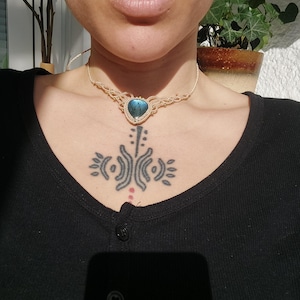 Pode incluir: Um colar de macramé bege com um pingente de pedra preciosa azul iridescente em forma de gota. O colar é usado por uma pessoa vestindo uma camisa preta. A imagem é um close-up, focando no colar e na parte superior do peito.