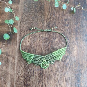 Puede incluir: Pulsera de macramé verde hecha a mano con cuentas doradas. La pulsera presenta un diseño tejido detallado y se muestra sobre una superficie de madera, con follaje verde en el fondo.