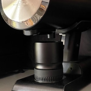 Puede incluir: Un molinillo de espresso negro con una parte superior plateada y el texto "WEBER" y "MM". También son visibles un pisón negro y un pequeño objeto redondo negro con un gráfico blanco. Los artículos están sobre una superficie oscura.