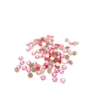 Puede incluir: Un conjunto de pedrería rosa con un reverso plano plateado. Los cristales redondos y facetados están dispersos sobre un fondo blanco, reflejando la luz. Estos adornos decorativos son adecuados para diversos proyectos de manualidades.